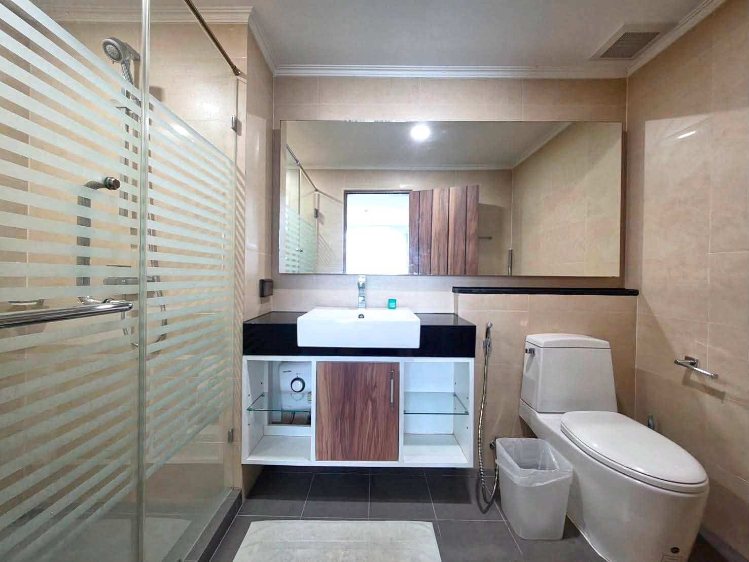 Dusit_1_2BR_Rent_Rdl279_11 Modern bathroom with a glass shower, white sink on a black countertop, large mirror, toilet, and beige tile walls.