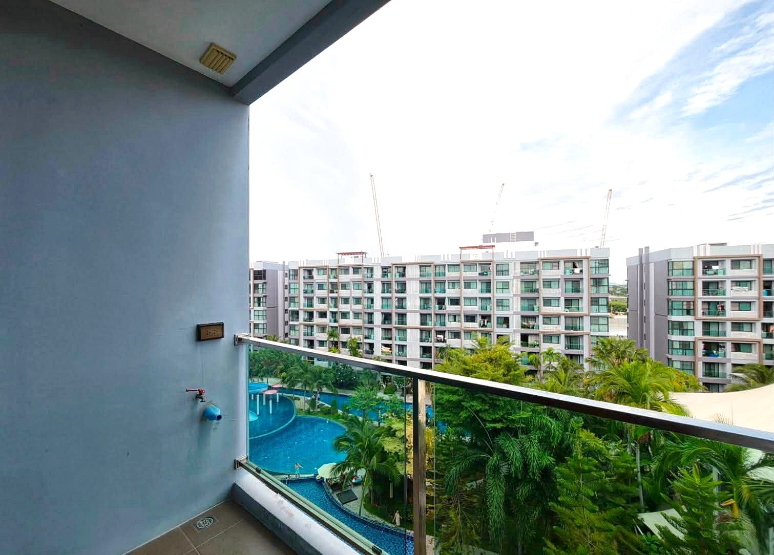 Dusit_1_2BR_Rent_Rdl279_15 Balcony view overlooking a pool area with palm trees and a row of modern white apartment buildings in the background.