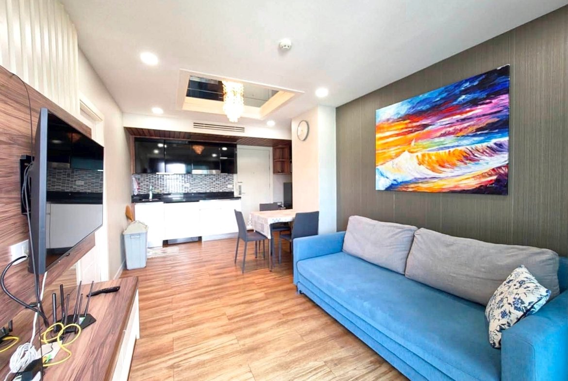 Open-concept living room with a blue sofa, abstract wall art, and a kitchen in the background.
