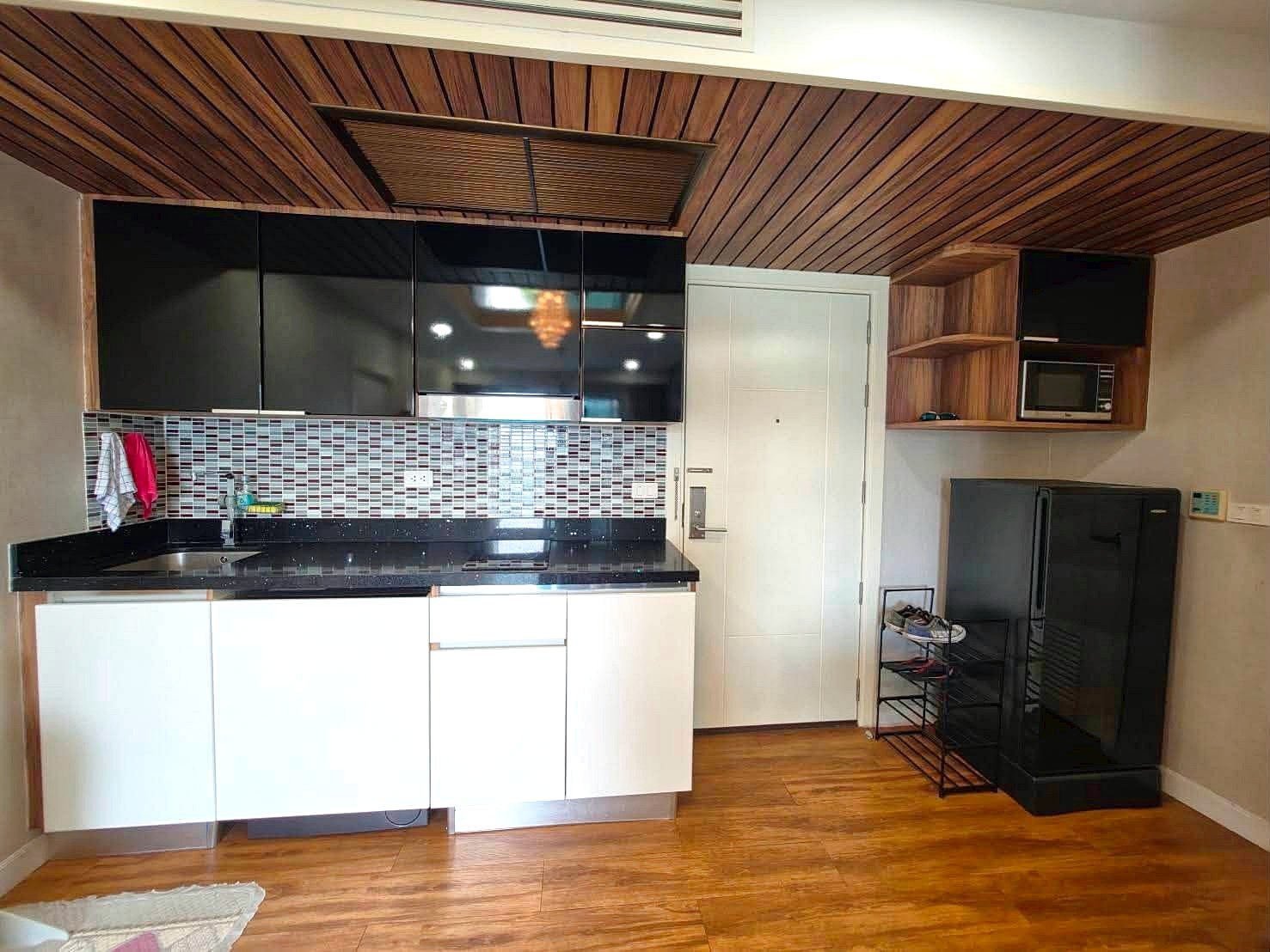 Dusit_1_2BR_Rent_Rdl279_4 Modern kitchen with black countertops, white cabinets, and a mosaic tile backsplash under a wooden slat ceiling.