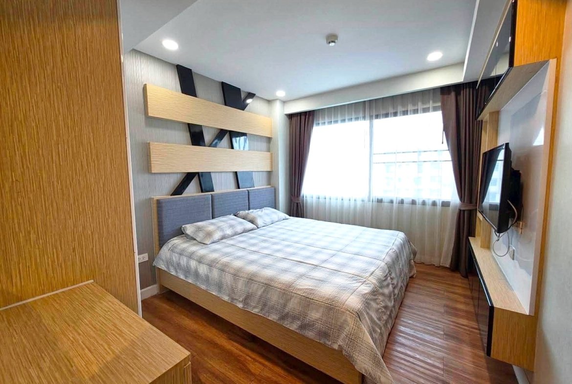 Modern hotel-style bedroom with a king bed, light plaid bedding, and a wall-mounted TV opposite the bed.