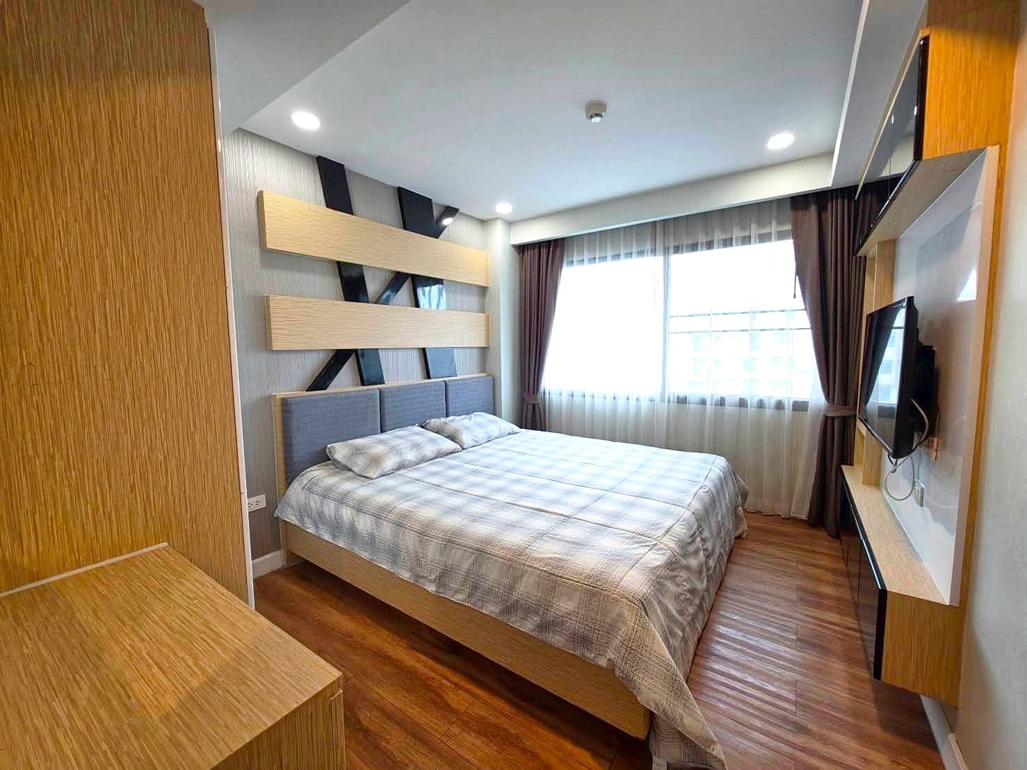 Dusit_1_2BR_Rent_Rdl279_5 Modern hotel-style bedroom with a king bed, light plaid bedding, and a wall-mounted TV opposite the bed.