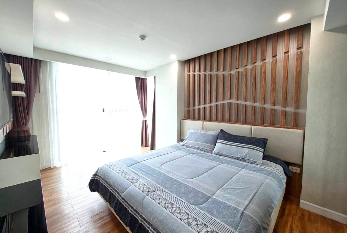 Hotel-style bedroom with a low platform bed, blue-gray quilt and pillows, against a vertical wooden slat headboard; large window with sheer white and brown curtains on the left.