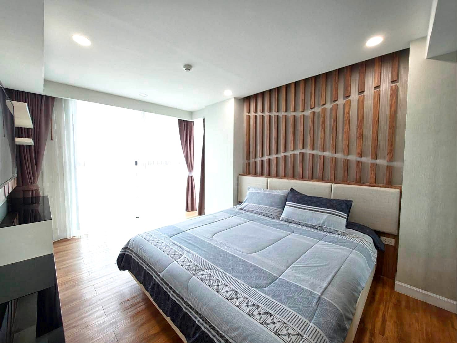 Dusit_1_2BR_Rent_Rdl279_9 Hotel-style bedroom with a low platform bed, blue-gray quilt and pillows, against a vertical wooden slat headboard; large window with sheer white and brown curtains on the left.