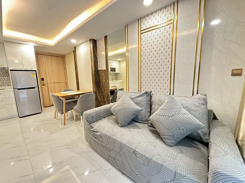 Open-plan living area featuring a gray patterned sofa with cushions, a wooden dining table, and a stainless steel fridge in a bright marble-floored space.