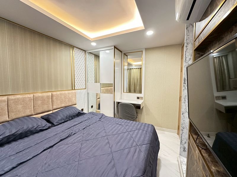 Modern hotel bedroom with a large navy quilted bed, padded headboard, mirrored wardrobes, a small desk with chair, and a wall-mounted TV.