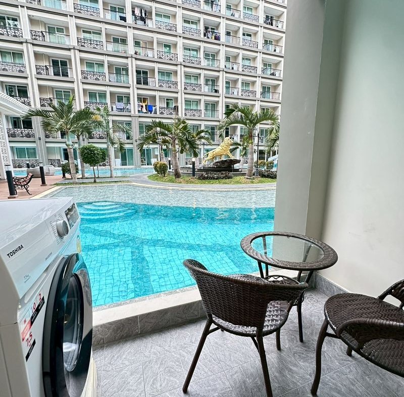 Balcony seating with a glass table and chairs overlooks a turquoise pool, palm trees, and a multi‑story apartment building; a washing machine is on the left edge.