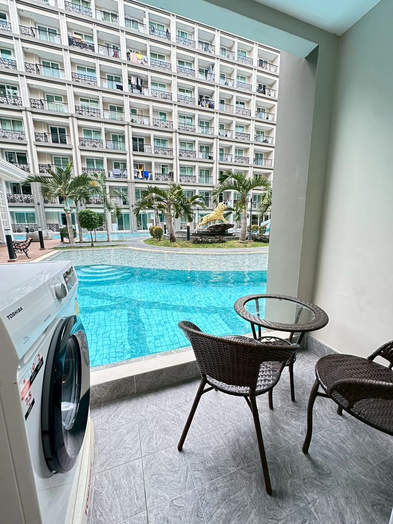 Dusit_2_1BR_Dlc556 4 Balcony seating with a glass table and chairs overlooks a turquoise pool, palm trees, and a multi‑story apartment building; a washing machine is on the left edge.