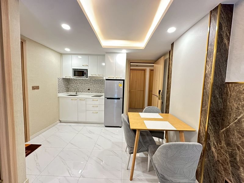 Open-plan kitchen with white cabinets, stainless steel fridge, and a wooden dining table with four upholstered chairs under recessed ceiling lights.