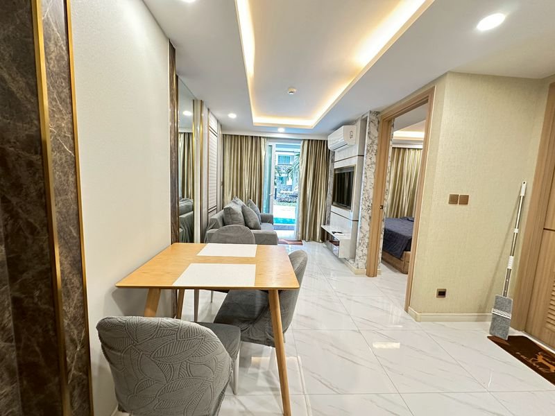 Open-plan living area with a wooden dining table and gray chairs, a gray sofa, a wall-mounted TV, and beige curtains over a balcony door.