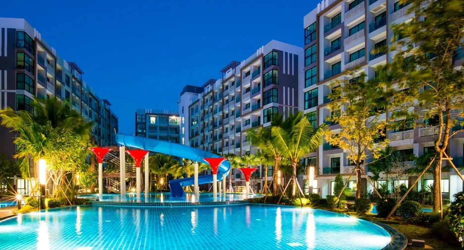 Resort-style apartment complex at dusk with a lit swimming pool and a blue water slide surrounded by palm trees and modern buildings.