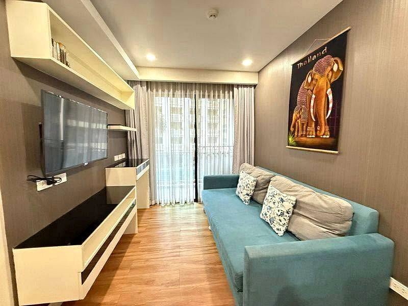 Cozy modern living room with a blue sofa and patterned cushions facing a wall-mounted TV and white entertainment unit; wooden floor, floor-to-ceiling sheer curtains, and an elephant artwork on the right wall.