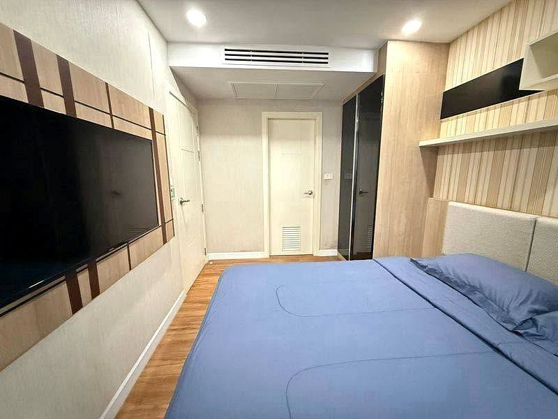 Modern hotel bedroom with a blue bedspread facing a wall-mounted TV; wood panel accents and a door at the far end.