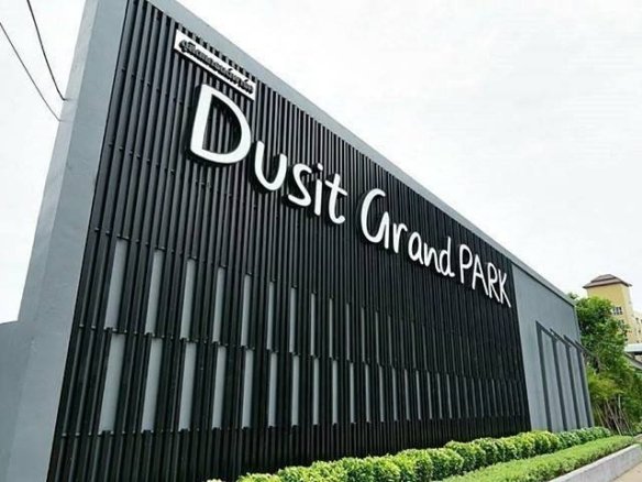 Exterior sign reading 'Dusit Grand Park' on a tall black panel wall with greenery at the base, angled street view.