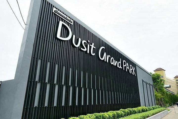 Exterior sign reading Dusit Grand Park on a tall black panel wall with greenery at the base angled street view