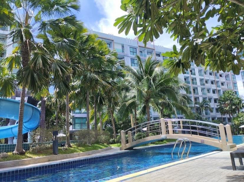 Resort pool area with a beige curved pedestrian bridge over a blue-tiled pool, palm trees, and a light hotel building in the background.