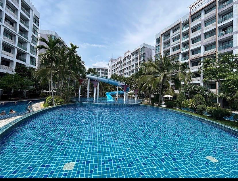 Curved blue-tiled pool at a resort with a blue water slide, surrounded by palm trees and white apartment buildings on both sides.