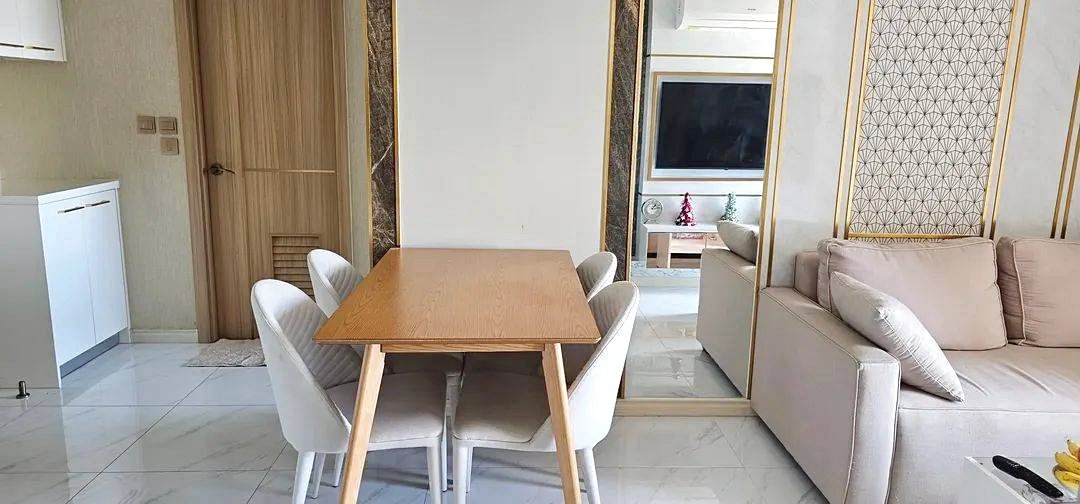 Rectangular wooden dining table with six light gray chairs in a modern open-plan living area; beige sofa on the right and a small Christmas tree visible beyond the doorway.