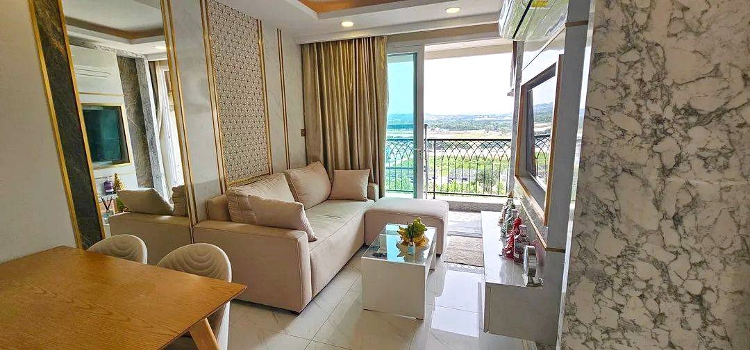 Bright living room with a beige sofa, glass coffee table, and a door to the balcony overlooking a distant landscape.