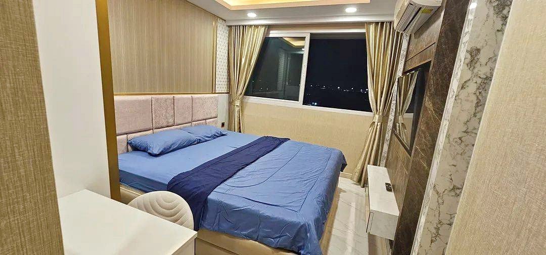 Hotel bedroom with a blue bedspread, beige headboard, and a large window showing city lights at night.