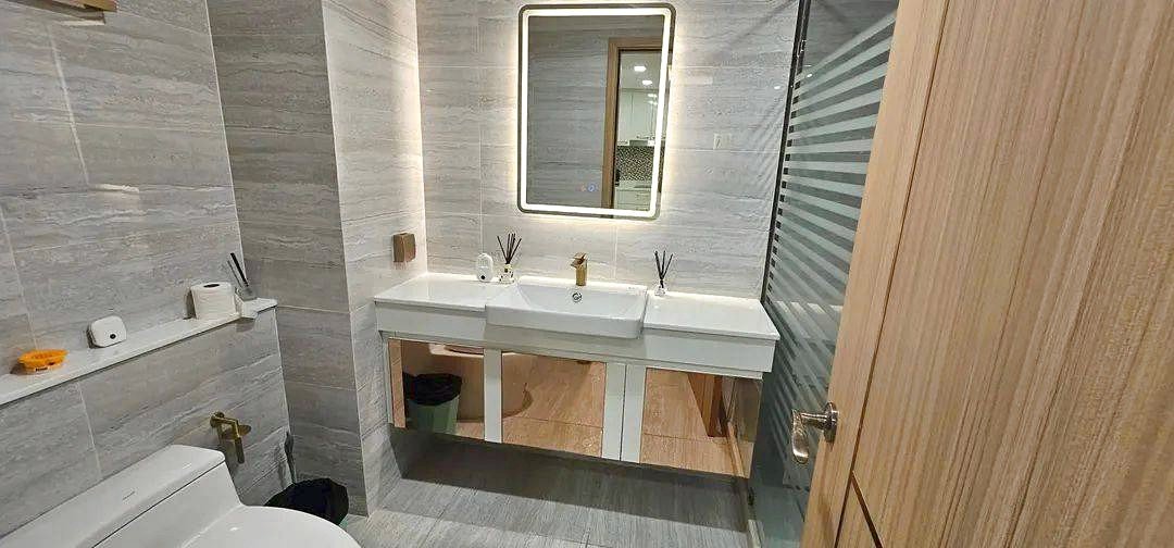 Modern bathroom with a double-sink vanity under a backlit square mirror, grey tile walls, and a glass shower door.
