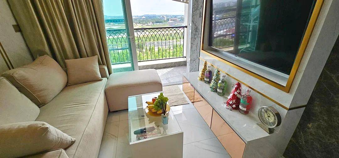 Bright living room with a beige sofa, glass coffee table, and a balcony view of greenery outside the window.