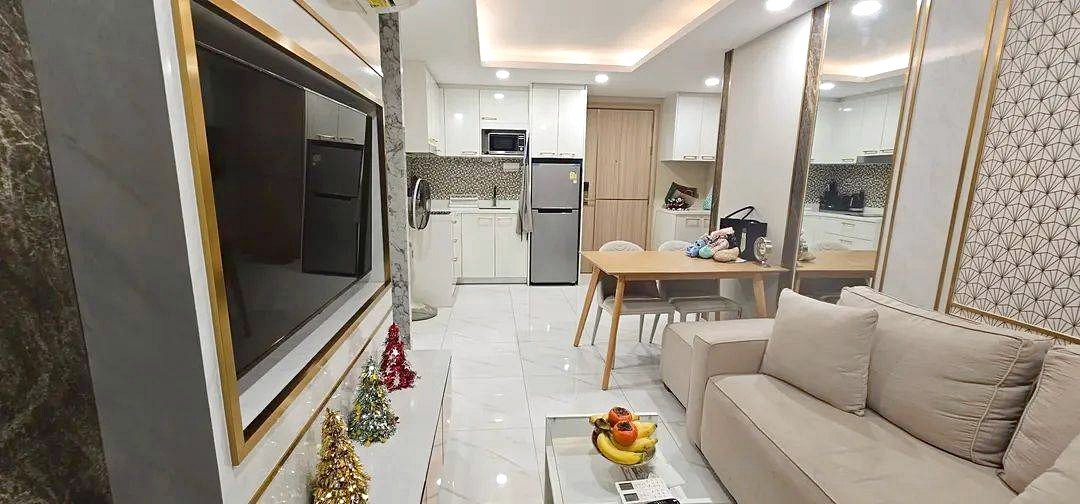 Open-concept living room with a large wall TV, beige sofa, glass coffee table, and a view into a white kitchen with stainless fridge; small Christmas trees on the TV stand.