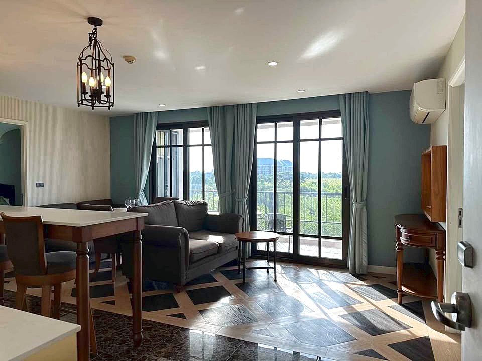 Sunlit living room with a gray sofa, round coffee table, and floor-to-ceiling glass doors opening to a balcony with a view of trees outside.