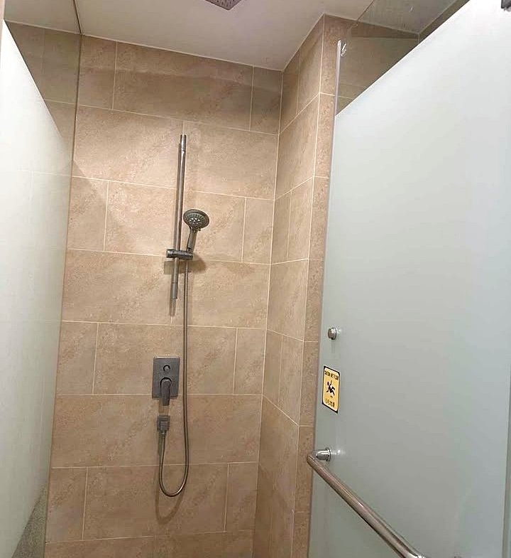 Shower stall with beige tiled walls, a ceiling rain shower head, and a handheld shower on a rail; frosted glass door with a chrome grab bar and safety sign.