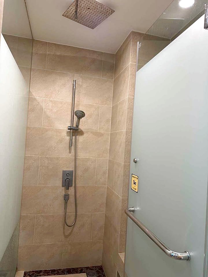 Espana_Condo_2BR_G517_518_11 Shower stall with beige tiled walls, a ceiling rain shower head, and a handheld shower on a rail; frosted glass door with a chrome grab bar and safety sign.