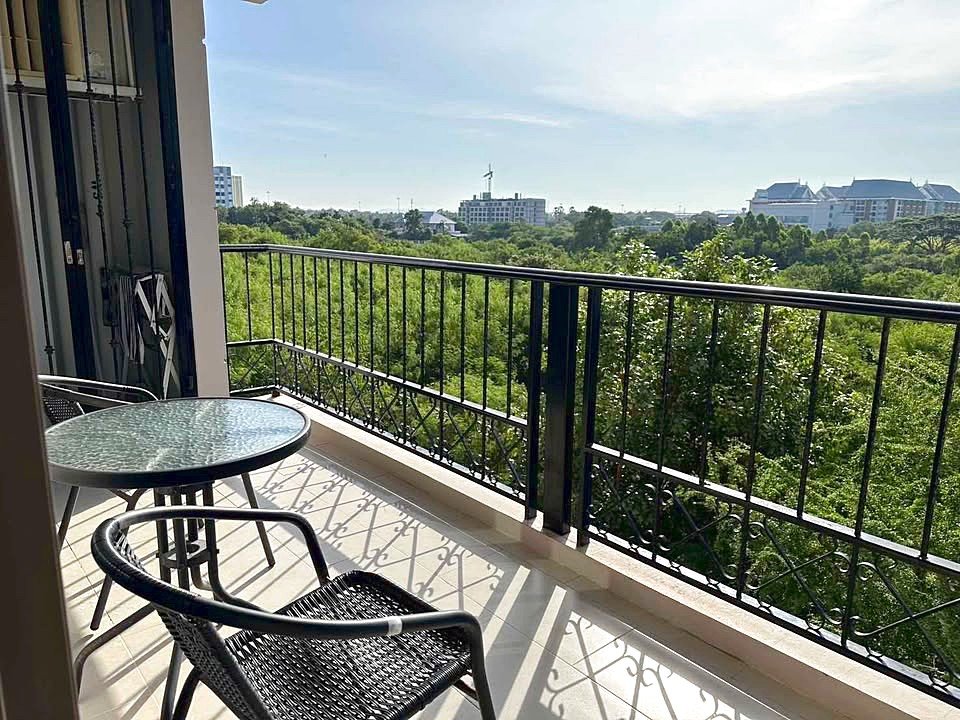 Balcony scene with a round glass table and two black wicker chairs, overlooking a green treetop view and distant buildings on a sunny day.