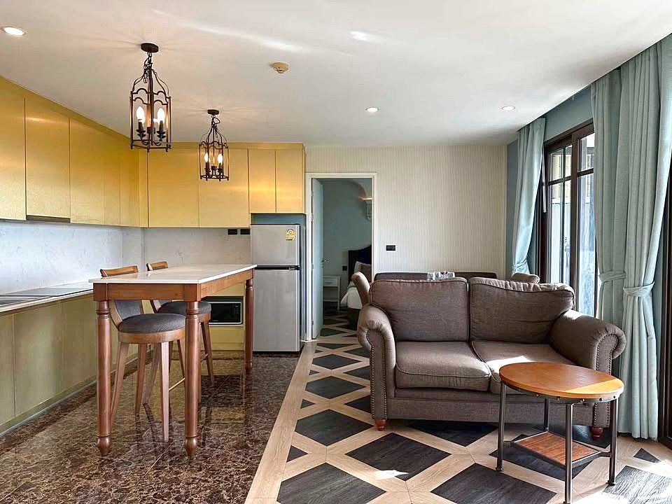 Open-concept living area with mustard-yellow kitchen cabinets, an island with two stools, and a brown sofa by a large window with teal curtains.