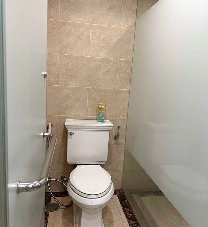 Toilet in a small tiled bathroom with a glass shower wall to the right and a chrome grab bar on the door side; a small blue bottle sits on the toilet tank.