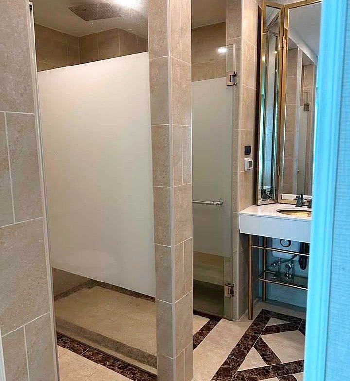Public bathroom interior with frosted shower stalls, beige tiles, and a vanity with mirror on the right.