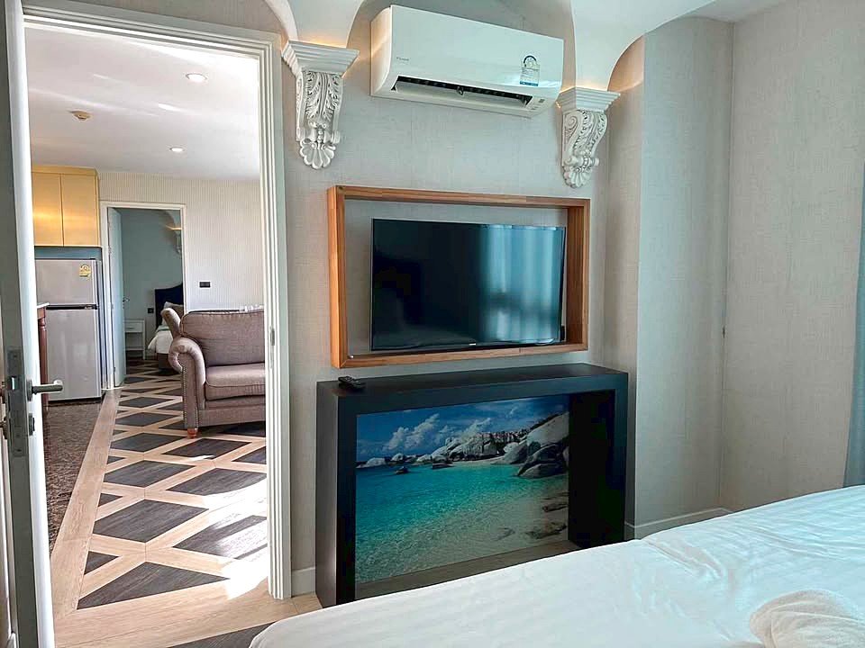 Hotel room interior with a wall-mounted TV above a console showing a beach image; doorway leads to a living area with a beige sofa and patterned floor. A ceiling air conditioner is mounted above.