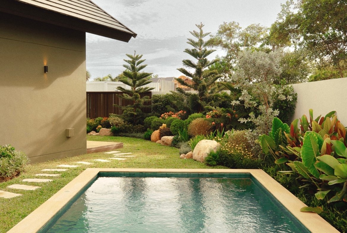 Rectangular backyard pool with turquoise water, beige coping, and a lush garden along the fence and wall of a beige house.