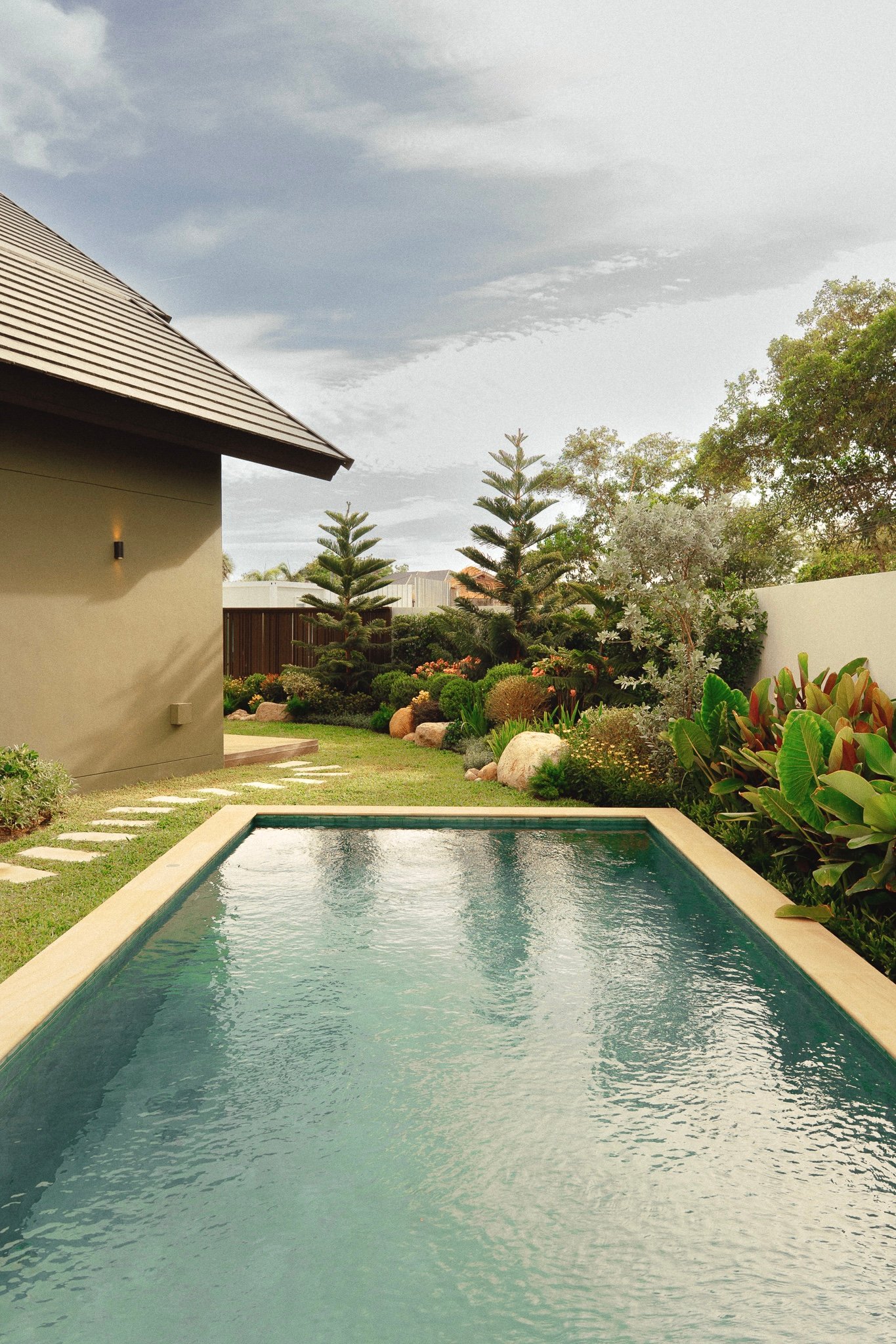 Rectangular backyard pool with turquoise water, beige coping, and a lush garden along the fence and wall of a beige house.