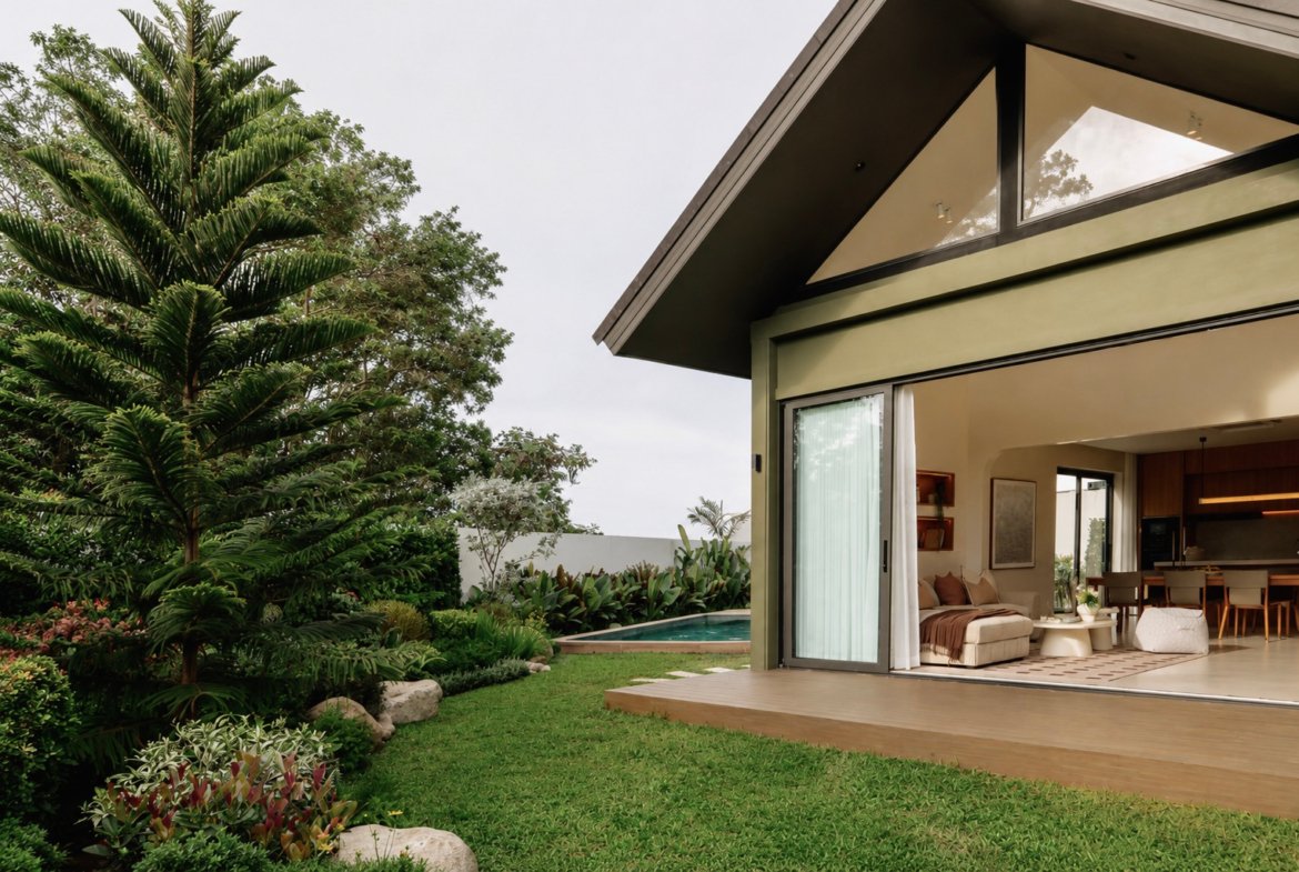 Open sliding glass doors reveal a modern living area with a sofa; a lush garden and pool sit just outside on a sunny day.