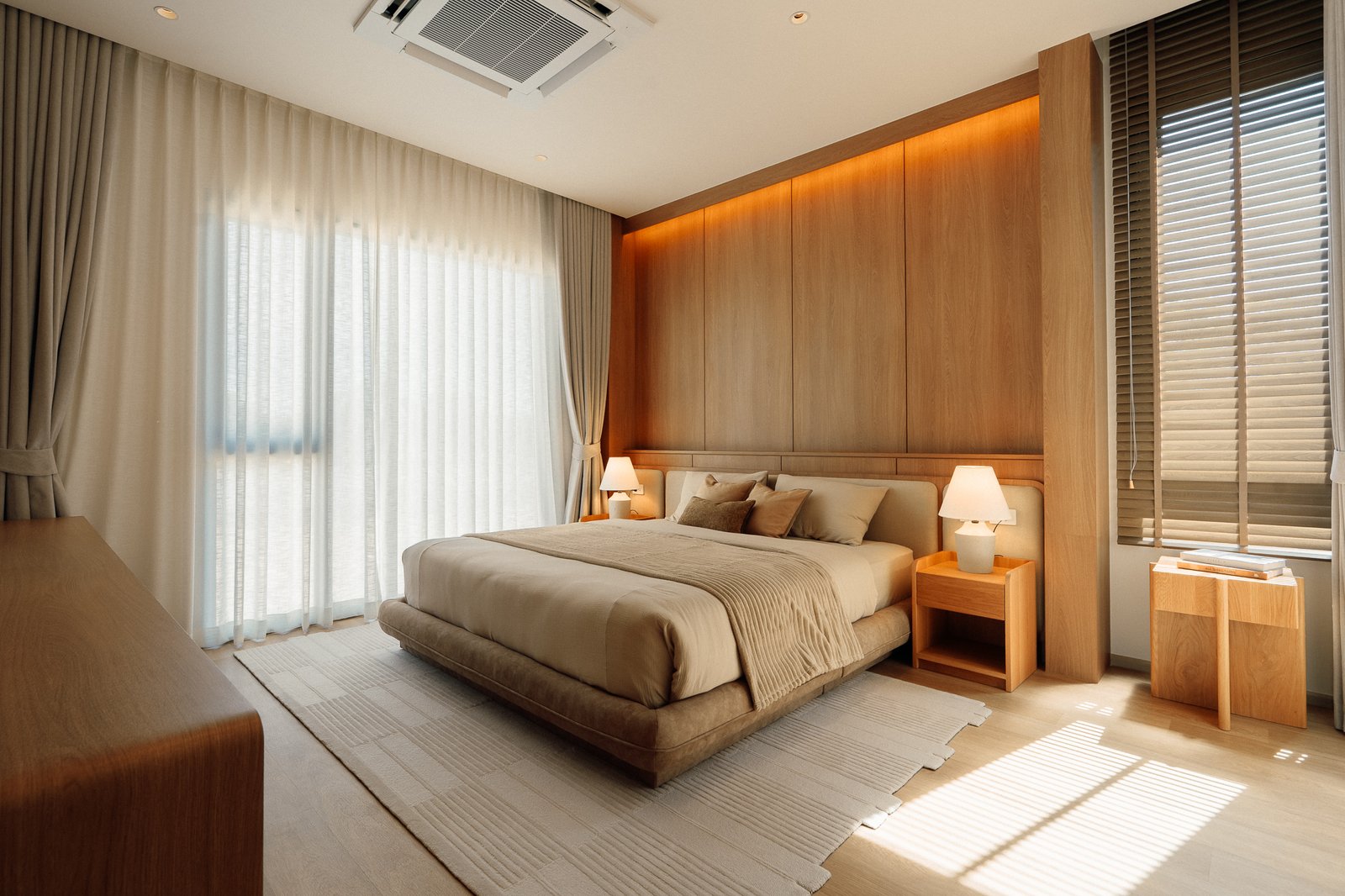 Sunlit hotel-style bedroom with a beige bed, sheer curtains, and a large wooden wardrobe along the wall