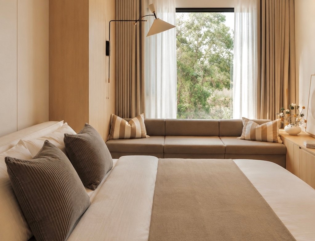 Hotel bedroom with a large bed, gray pillows, and a beige sofa by a window with sheer curtains.
