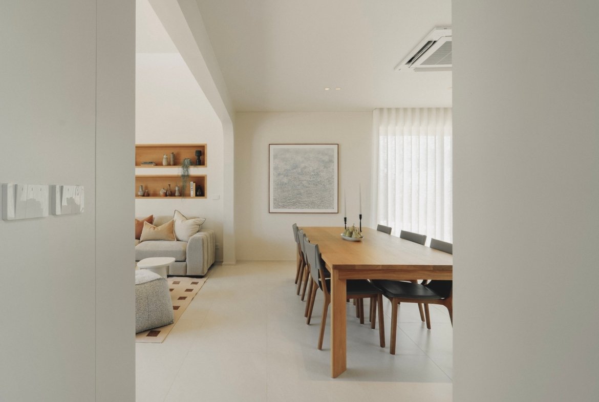 Open-plan dining area with a wooden table and chairs, a beige sofa, and built-in wall shelves in a bright, neutral living space.