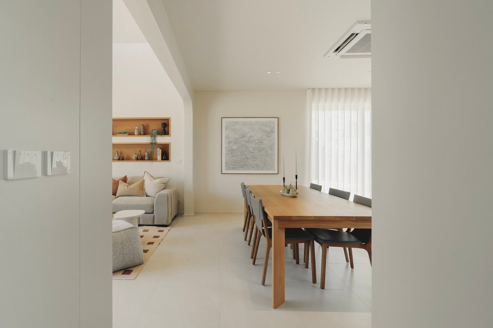 Open-plan dining area with a wooden table and chairs, a beige sofa, and built-in wall shelves in a bright, neutral living space.