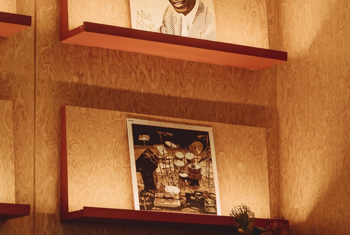 Wood-paneled wall with red floating shelves showing a Nat King Cole portrait and a second framed photo below (drum set scene).