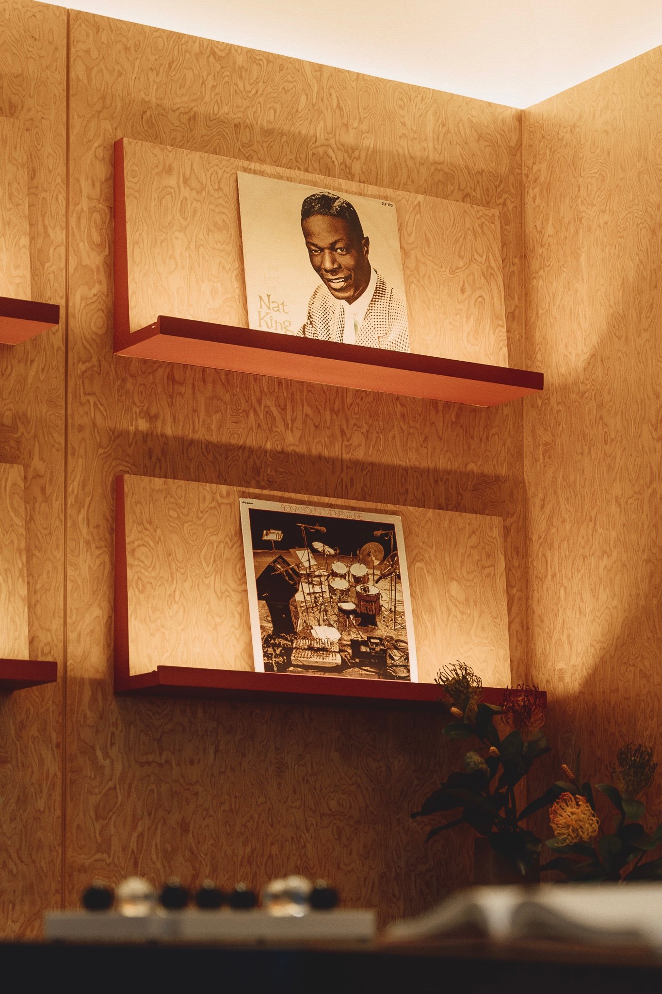 Wood-paneled wall with red floating shelves showing a Nat King Cole portrait and a second framed photo below (drum set scene).