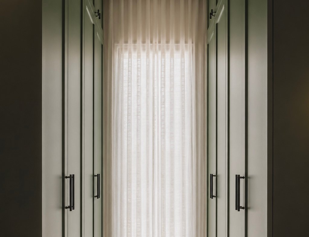 Narrow walk‑in closet with tall green paneled wardrobes on both sides and a sheer white curtain at the end letting light in.