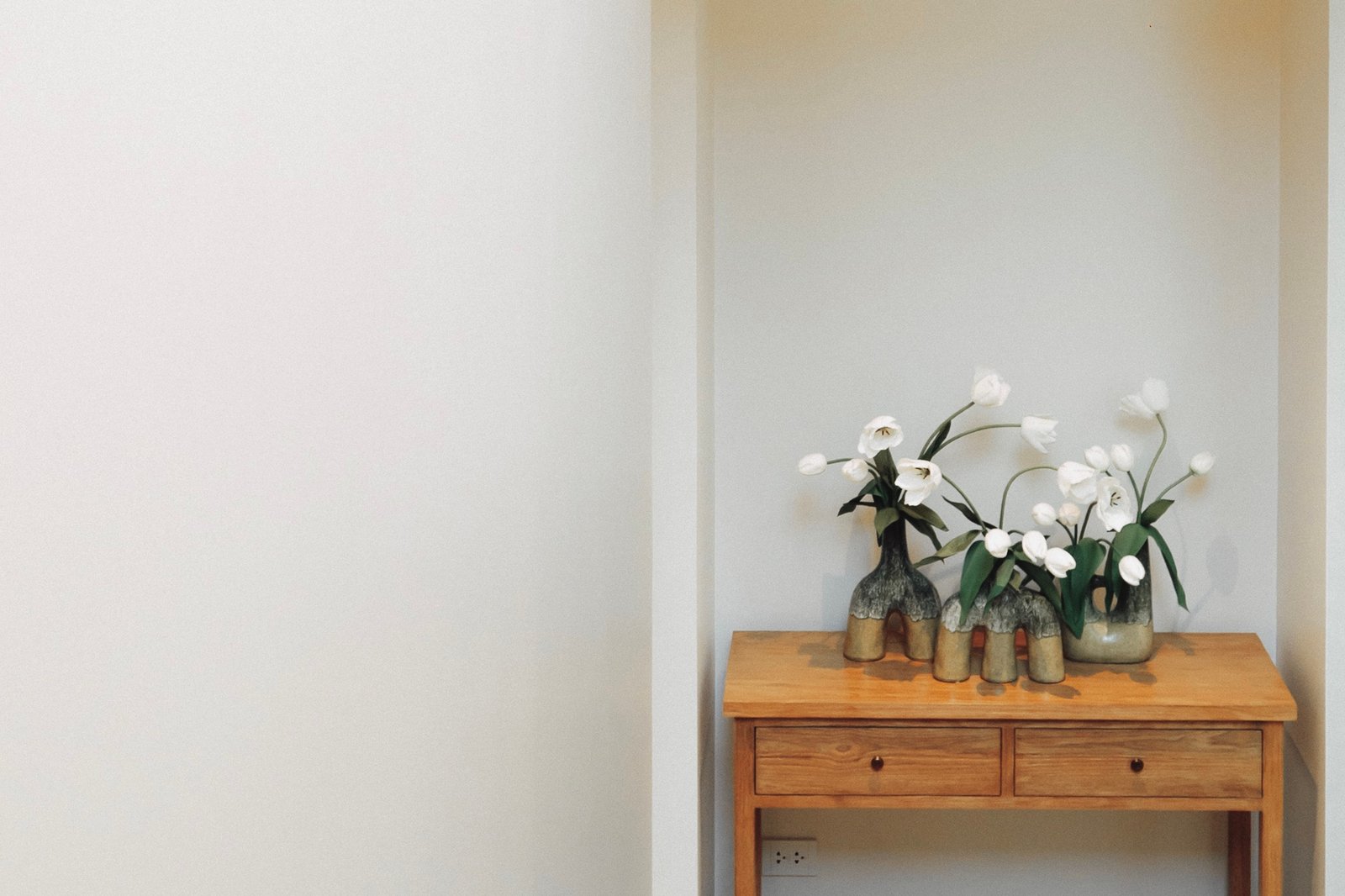 Wooden dresser topped with three ceramic vases of white tulips against a pale wall.
