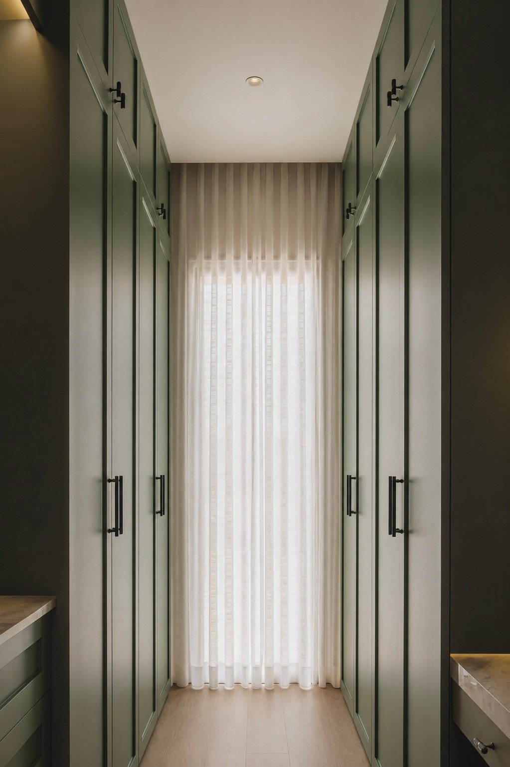 Narrow walk‑in closet with tall green paneled wardrobes on both sides and a sheer white curtain at the end letting light in.