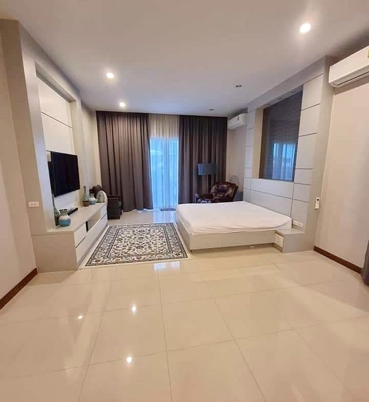 Modern hotel-style bedroom with white furniture, low platform bed, wall TV, and a seating area by large windows with dark curtains and a patterned rug.