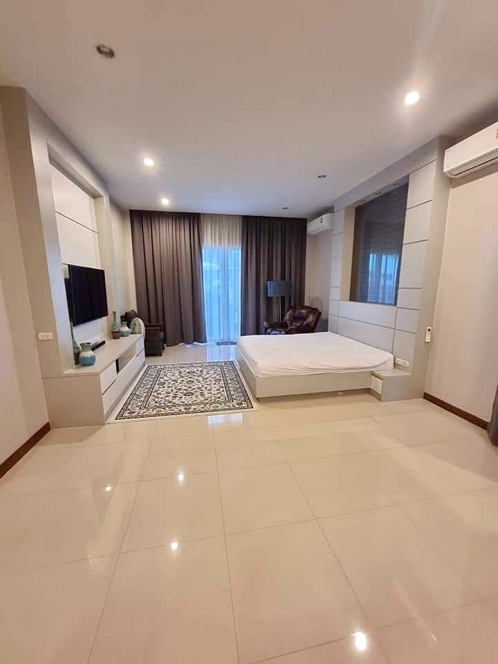 Garden_Ville_1_Pattaya_Rent_Rdl368_10 Modern hotel-style bedroom with white furniture, low platform bed, wall TV, and a seating area by large windows with dark curtains and a patterned rug.