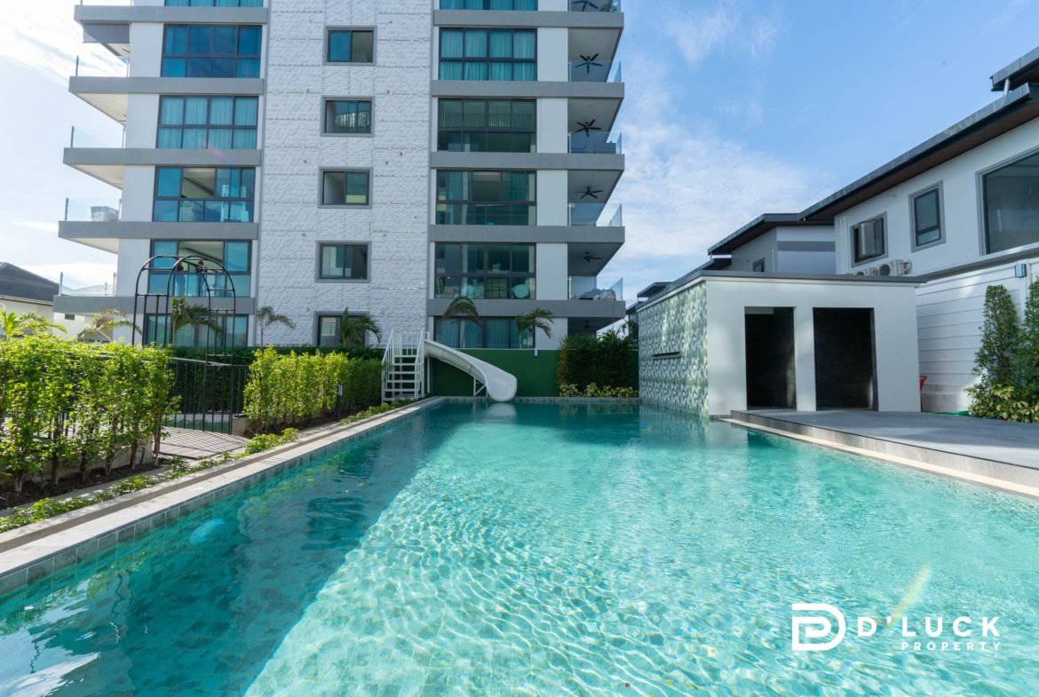 Modern white apartment building with glass balconies beside a long turquoise pool under a blue sky. Water slide and greenery at pool edge visible.